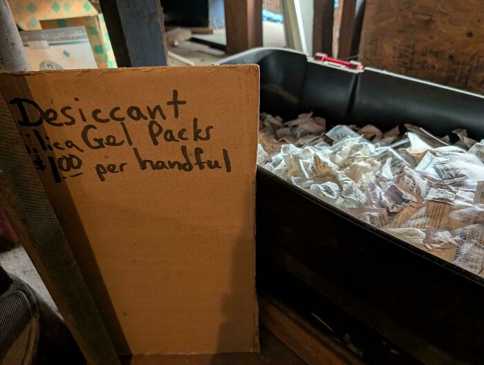 Cardboard sign at thrift store selling handfuls of silica gel desiccant packs for one dollar in a large black bin.