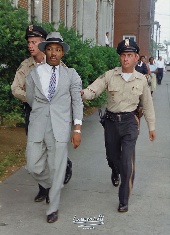 Martin Luther King Jr. being escorted by police officers in an interesting colorized history pic on a city sidewalk.