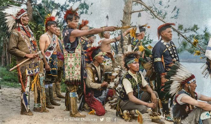 Colorized history pic of Native American men in traditional regalia participating in a cultural ceremony outdoors.