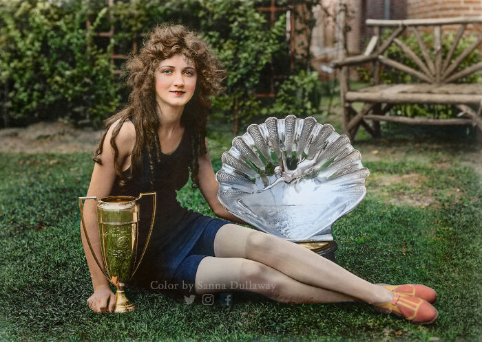 Young woman sitting on grass with large trophies in an interesting colorized history pics photograph.
