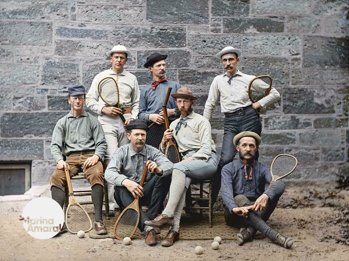 Colorized history pic of early tennis players wearing vintage clothing and holding wooden rackets posing outdoors.