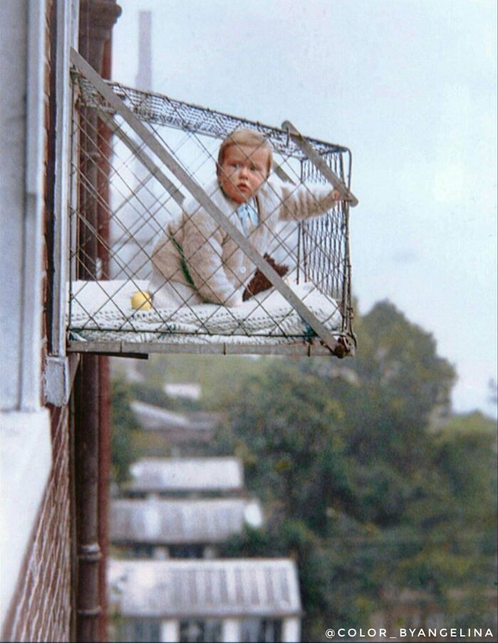 Child in a metal cage balcony outside a building, a rare interesting colorized history pic showing past child safety measures.