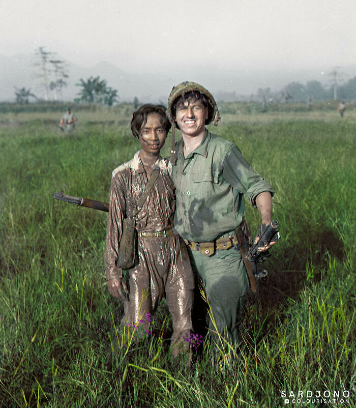 Two soldiers, one muddy and one in uniform, smiling together in a grassy field, an interesting colorized history pic.
