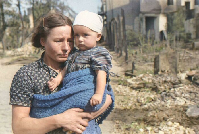 Woman holding baby wrapped in a blue blanket stands in ruined street, an interesting colorized history pics moment from war era.