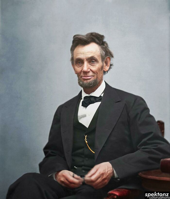 Colorized history pic of Abraham Lincoln seated, wearing a black suit and bow tie, against a muted gray background.