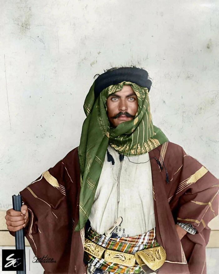 Colorized history image of a traditional man wearing a green headscarf and brown robe holding a long object.