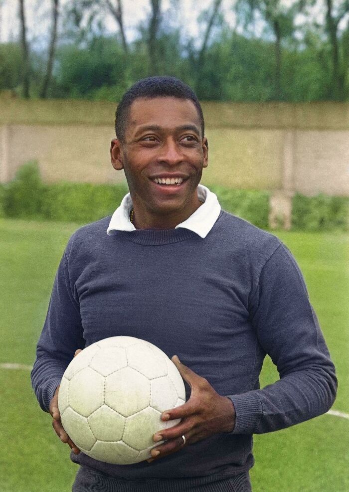 Colorized history pic of a smiling man holding a soccer ball on a grassy field with trees in the background.