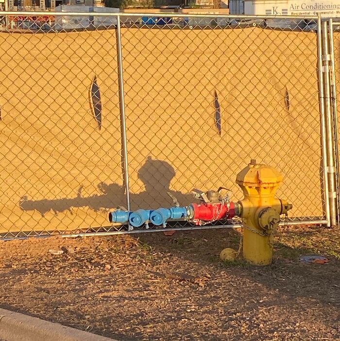 Shadow of fire hydrant and pipes creating an interesting and bizarre silhouette on a fenced wall in bright sunlight.