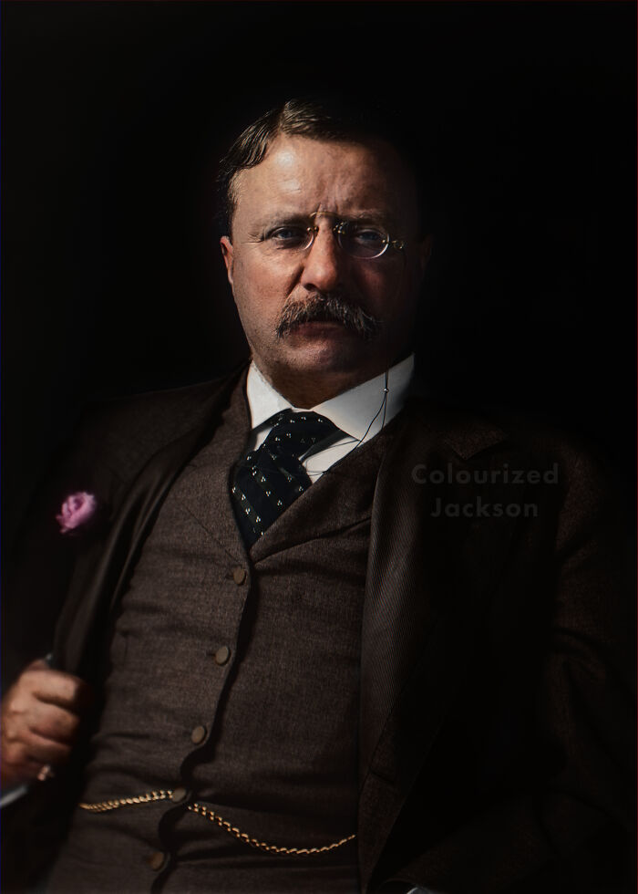 Colorized history pic of a serious man with glasses and mustache wearing a suit and tie against a dark background.
