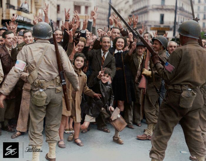 Colorized history photo of World War II soldiers greeting joyful civilians in a lively street scene with smiles and peace signs.