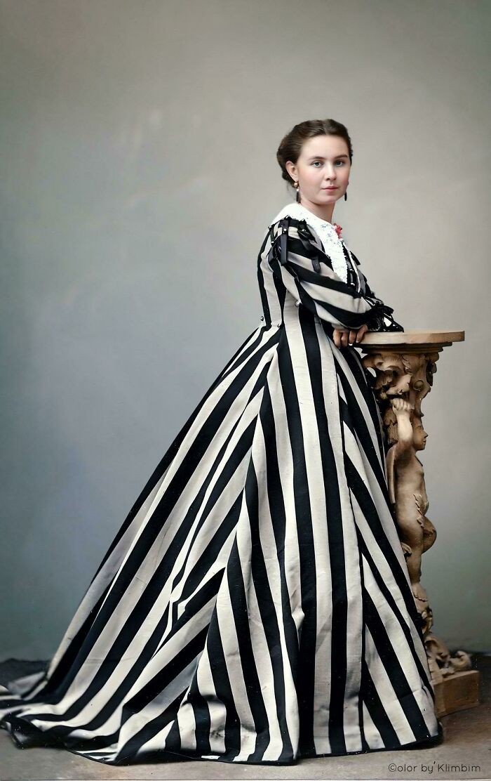 Young woman in a colorized historical portrait wearing a black and white striped Victorian-era dress standing by a carved pedestal.