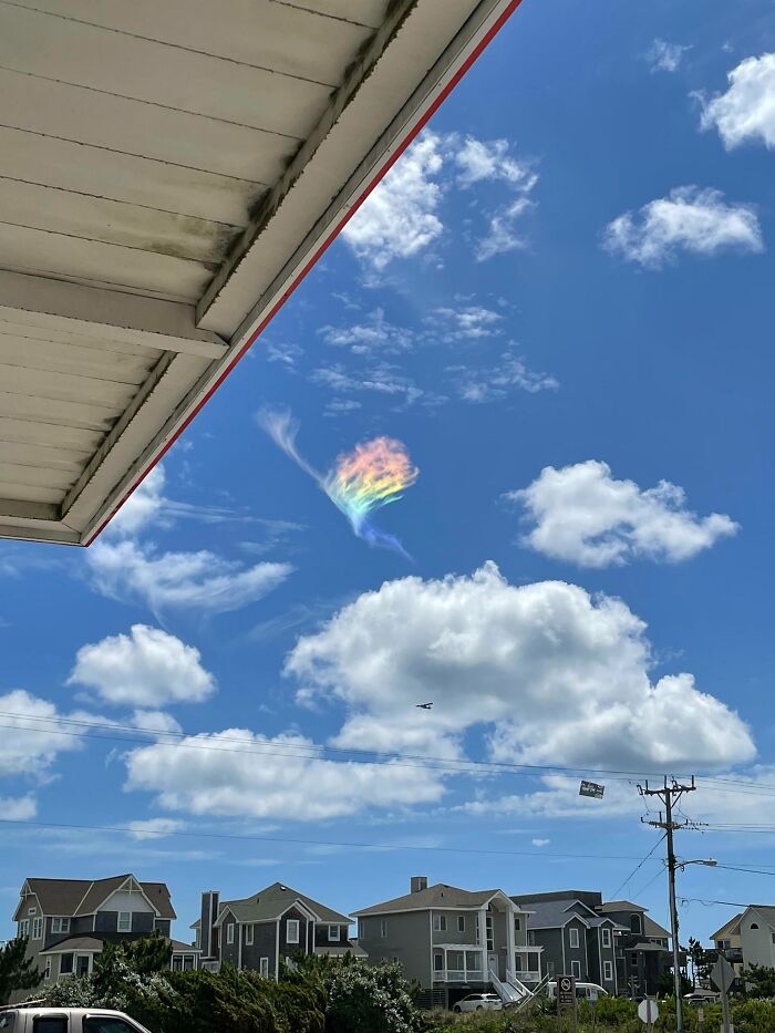 Unreal photograph of a rare rainbow cloud formation above suburban houses under a bright blue sky with scattered clouds.