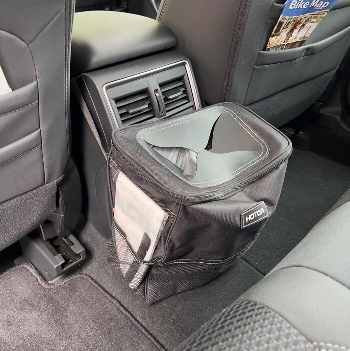 Black portable car trash bag with tissue dispenser placed on the floor behind the front seats, showcasing weirdly wonderful items.
