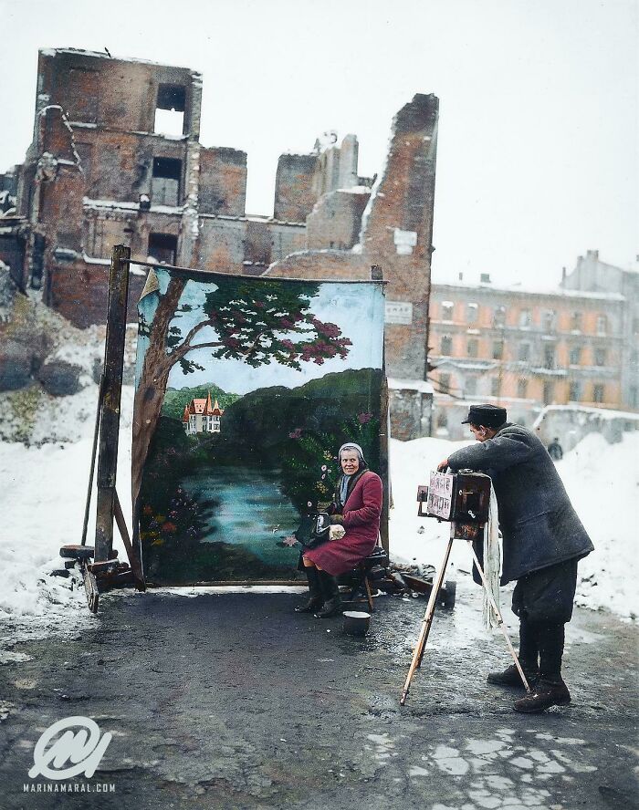 Colorized history pic of a woman posing for a photo with a painted scenic backdrop in front of war-damaged buildings.