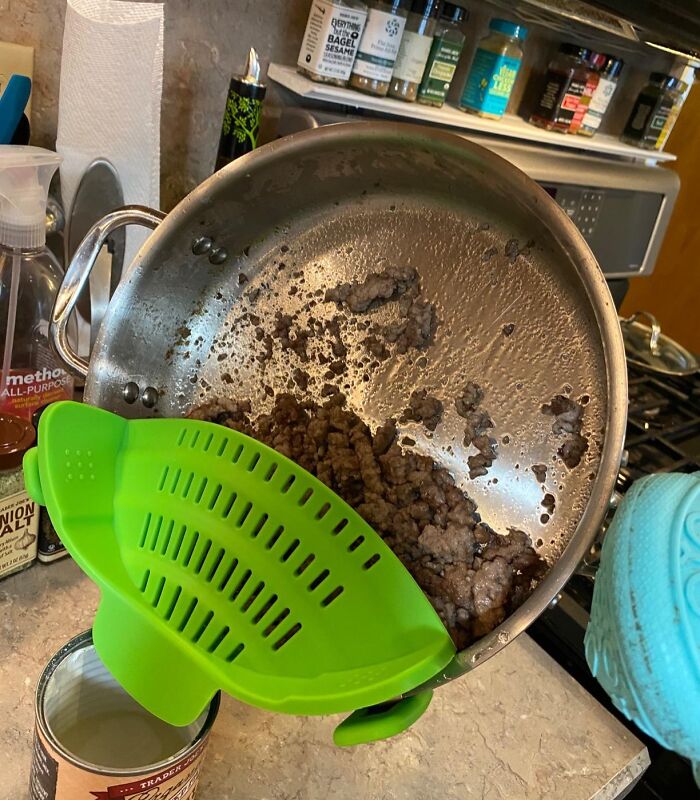 Green silicone strainer attached to old cooking pan draining grease from browned ground meat in kitchen setting.