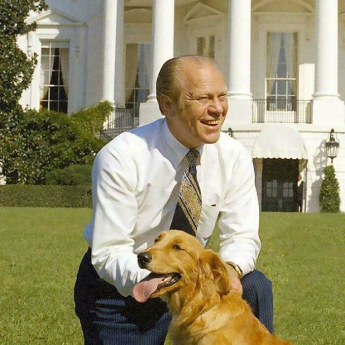 Man in white shirt and tie smiling outside a large building, sitting on grass with a happy golden retriever dog.