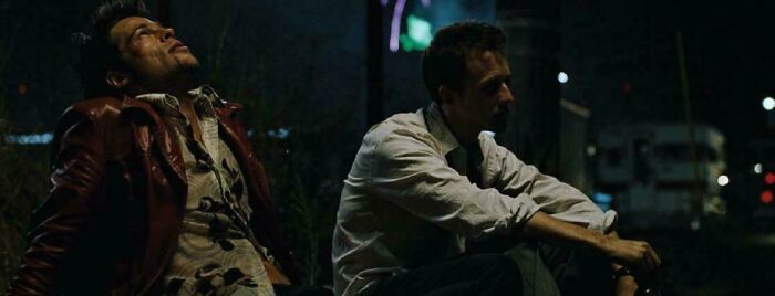 Two men sitting outdoors at night, one leaning back with eyes closed, the other looking ahead thoughtfully.