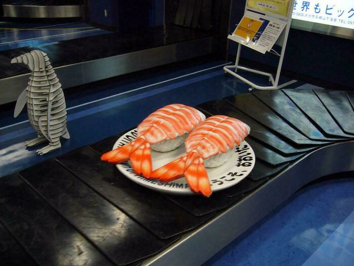 Two large shrimp sushi models on a conveyor belt with a wooden penguin figure nearby, fascinating content display.