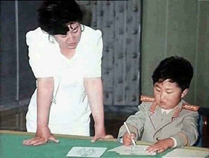 A child in a military uniform writing at a table while a woman in white watches, illustrating fascinating content on the internet.