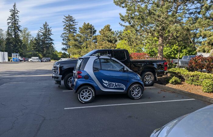 Small Hot Wheels branded car parked next to large black pickup trucks, illustrating size comparison and variety.