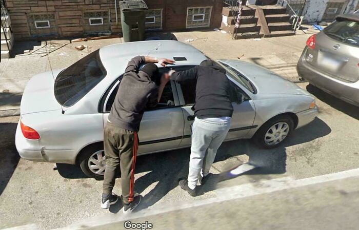 Two people caught in a shenanigan leaning into a car on a street, captured on Google Maps.