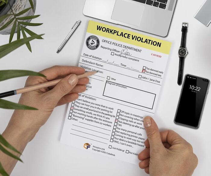 Person filling out a workplace violation form at a desk with laptop, phone, watch, and pen, office essentials visible