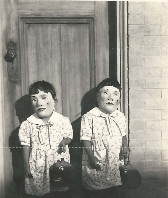 Two children in vintage creepy masks and dresses holding Halloween pumpkin buckets in front of an old wooden door.