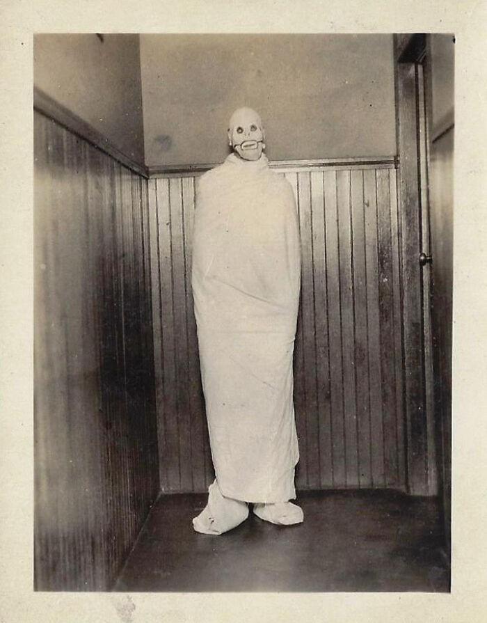 Black and white vintage photo of a figure wrapped in cloth with a creepy mask standing in a corner, unsettling past image.