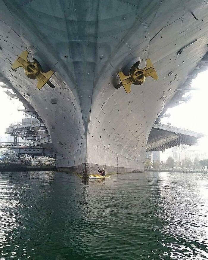 Kayaker paddling under a massive ship hull, showcasing fascinating comparison pics that highlight variety and scale differences.
