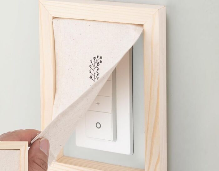 Hand covering light switch with fabric and wooden frame as a sneaky way to hide ugly eyesores in a home.