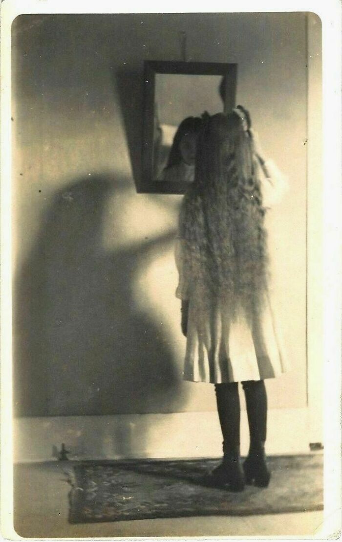Vintage eerie photo of a girl with long hair facing a mirror, casting a creepy shadow on the wall, unsettling and haunting.