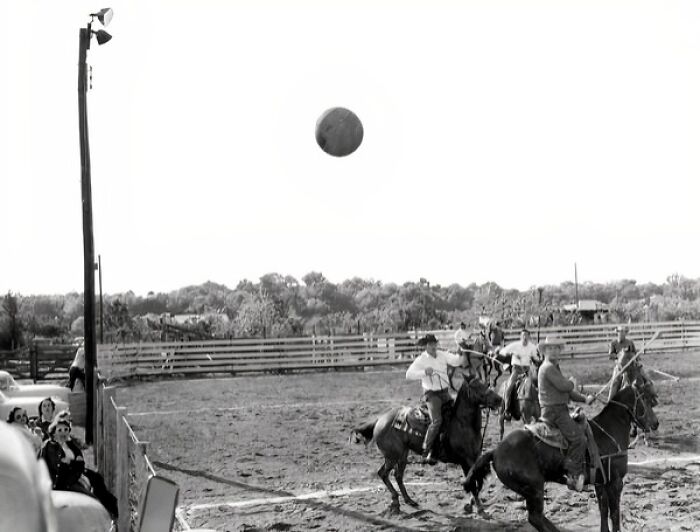 Vintage cowboy photograph showing men on horseback playing ball in a dusty outdoor arena with spectators watching.