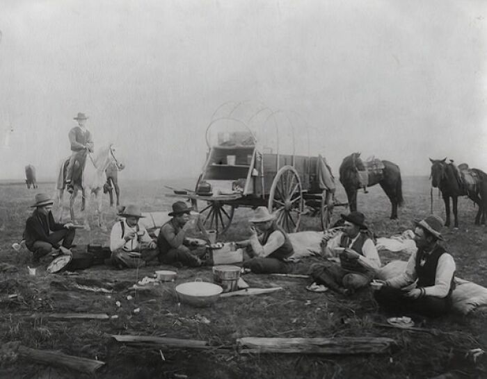 Group of vintage cowboys eating by a wagon and horses in an open field during the era when the West was wild.