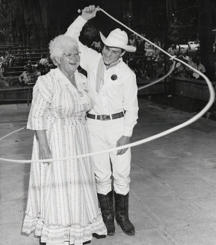 Young cowboy demonstrating lasso skills next to an elderly woman in a vintage cowboy photograph from the wild west era.