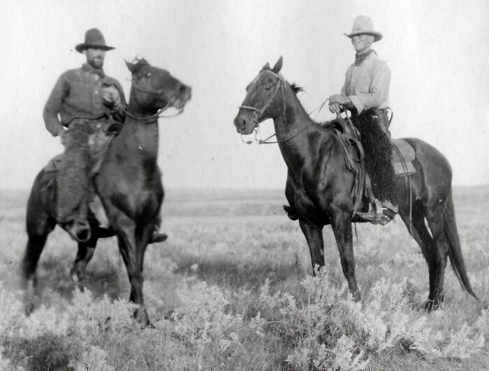 Two vintage cowboys on horseback in an open field, showcasing classic cowboy attire and Western lifestyle.