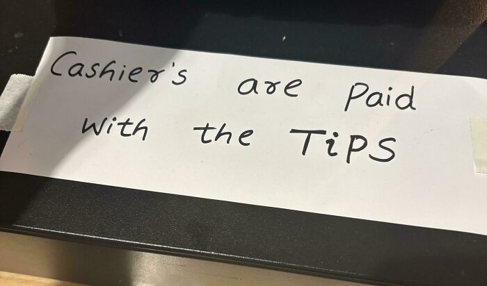Handwritten sign on white paper stating cashiers are paid with the tips, highlighting tipping culture issues.