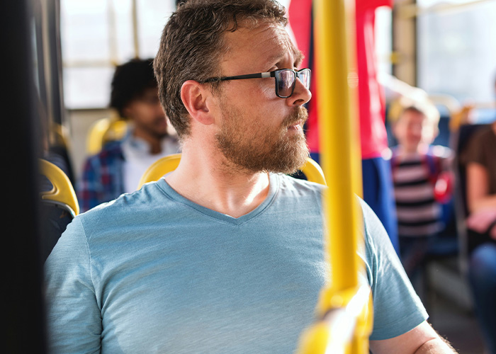 Man wearing glasses looking out window while sitting on crowded public transport during daytime ride.