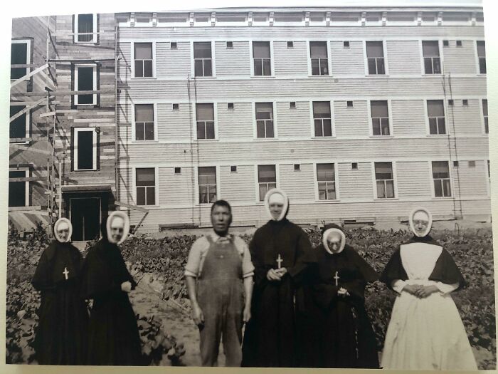 Vintage photo of nuns and a man in overalls standing in front of a large building, evoking creepy and unsettling vibes.