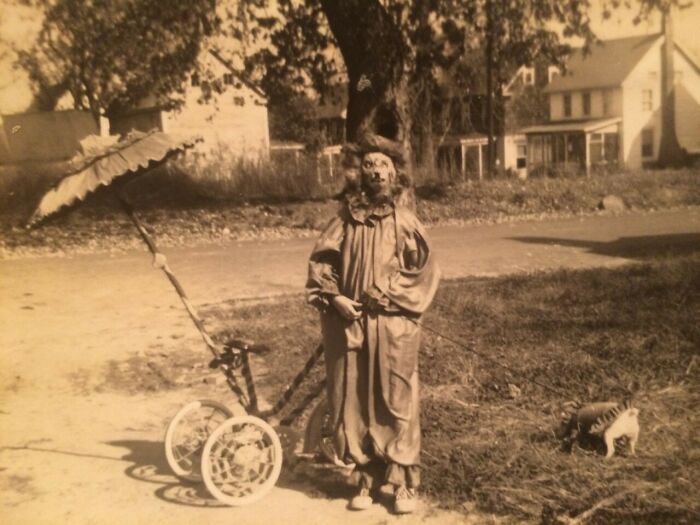 Sepia-toned creepy vintage photo of a child clown standing next to a tricycle and a small dog on a quiet street.