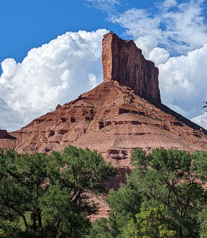 Red rock formation captured from an unexpected angle with green trees in foreground under a blue sky with clouds.