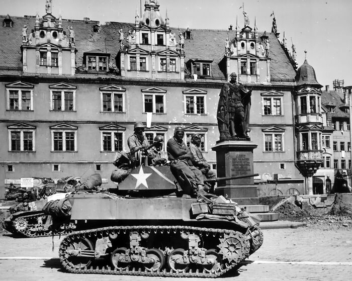 African American soldiers riding a military tank in a historic European town square during World War II.