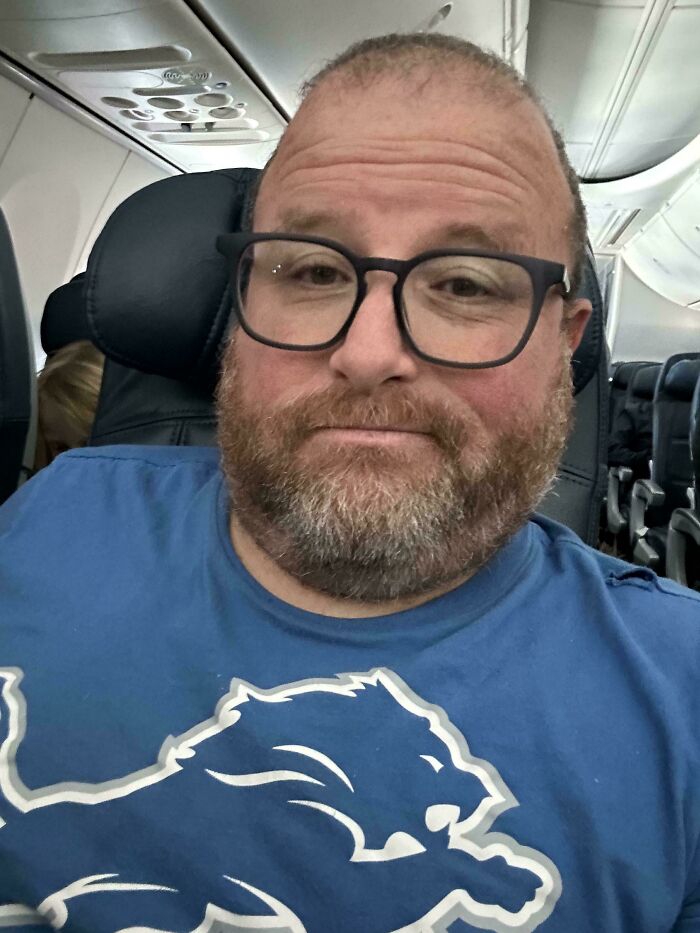Middle-aged man wearing glasses and a blue lion shirt sitting in an airplane seat conquering fear of flying.