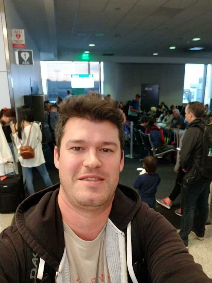 Man taking a selfie at a busy airport terminal, surrounded by travelers facing fear of flying and boarding planes.