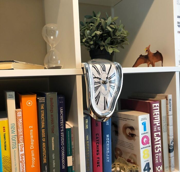 Melting clock on bookshelf surrounded by quirky gems like books, hourglass, plant, and toy dinosaur brightening the day.