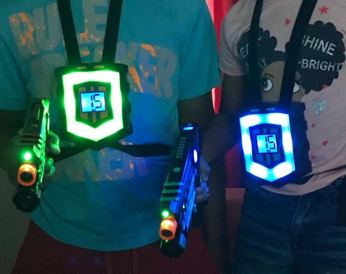 Two kids wearing light-up vests and holding toy laser guns in a dark room, perfect for shopping for a 12-year-old gift.