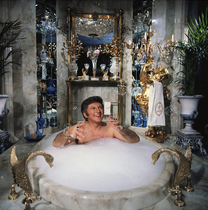 Man enjoying bubble bath in extravagant vintage bathroom with gold decor and mirrors, a ridiculous pic from the past.