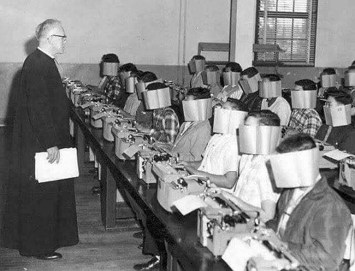 Black and white photo of students with face shields typing on typewriters in a vintage classroom setting, a ridiculous pic from the past.
