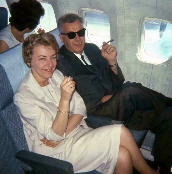 Couple smoking cigars relaxed on airplane seats in a vintage photo showing past habits that would never fly today.