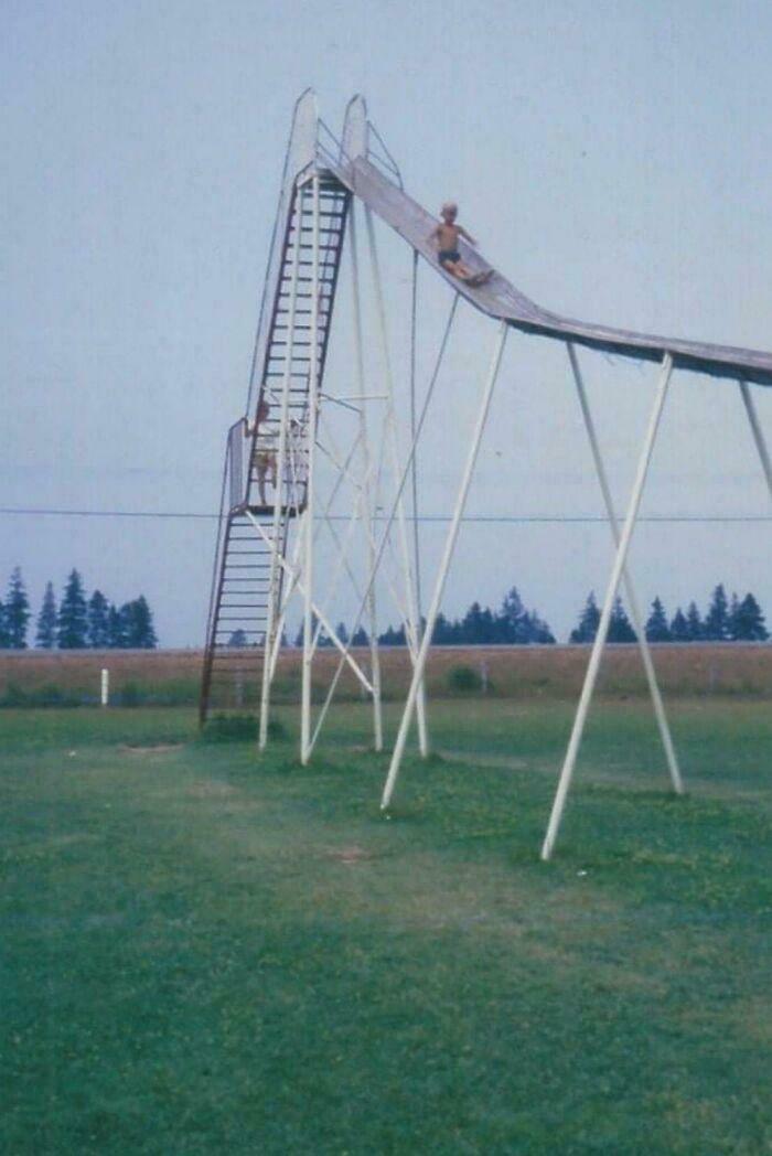 Child sliding down a tall vintage playground slide in a grassy field, one of the ridiculous pics from the past.
