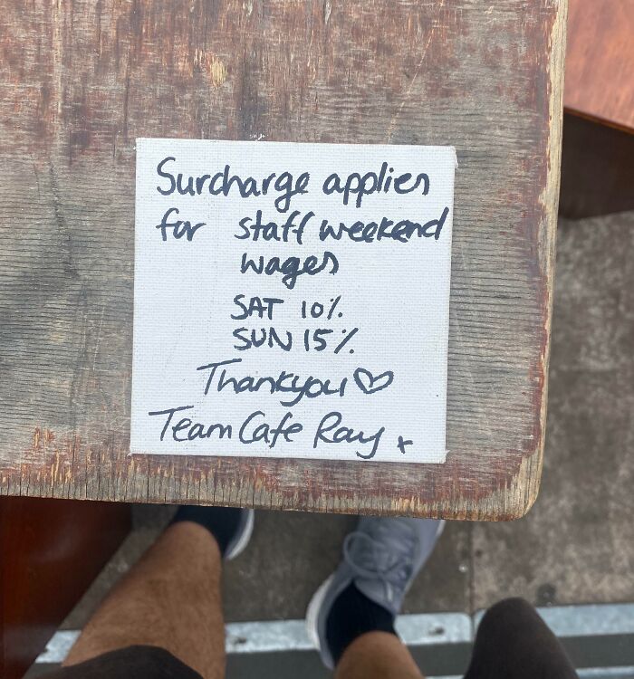 Handwritten note on wooden table explaining weekend surcharge for staff wages, highlighting ridiculous tipping culture.